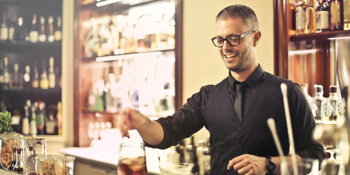 Happy young male barkeeper standing at counter and preparing alcohol cocktail for order while working in modern pub