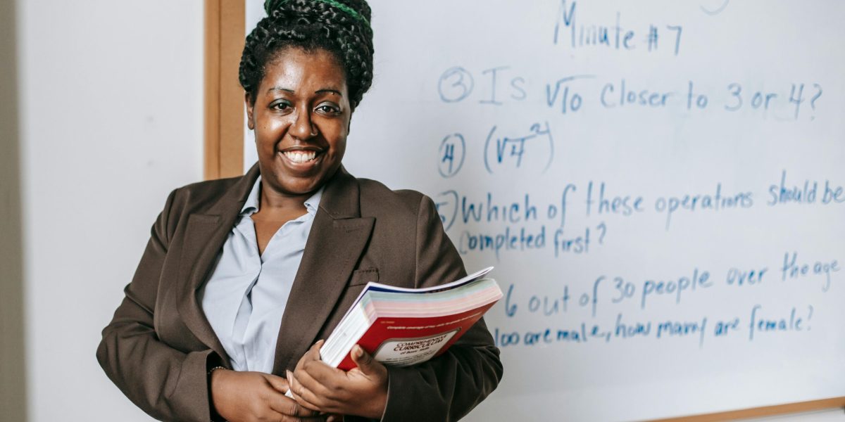 Cheerful black female teacher with workbooks standing near whiteboard