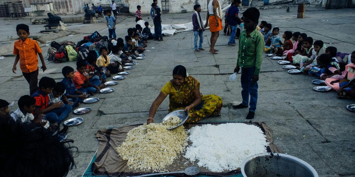 From above of old Indian woman serving food on plates while sitting on ground surrounded with kids in poor area